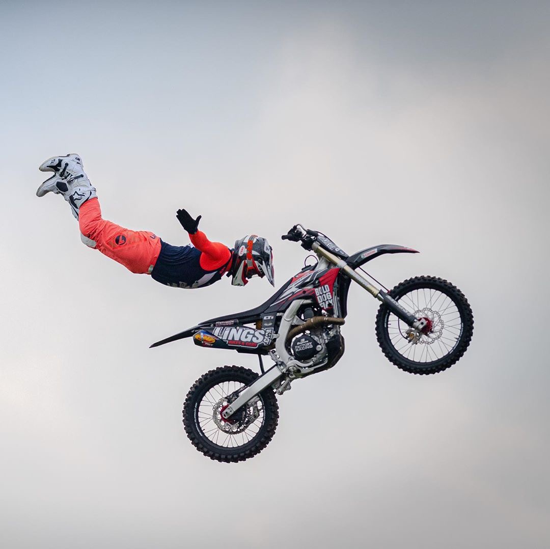 Piloto de motocross freestyle em pleno salto, com o corpo estendido no ar ao lado da moto